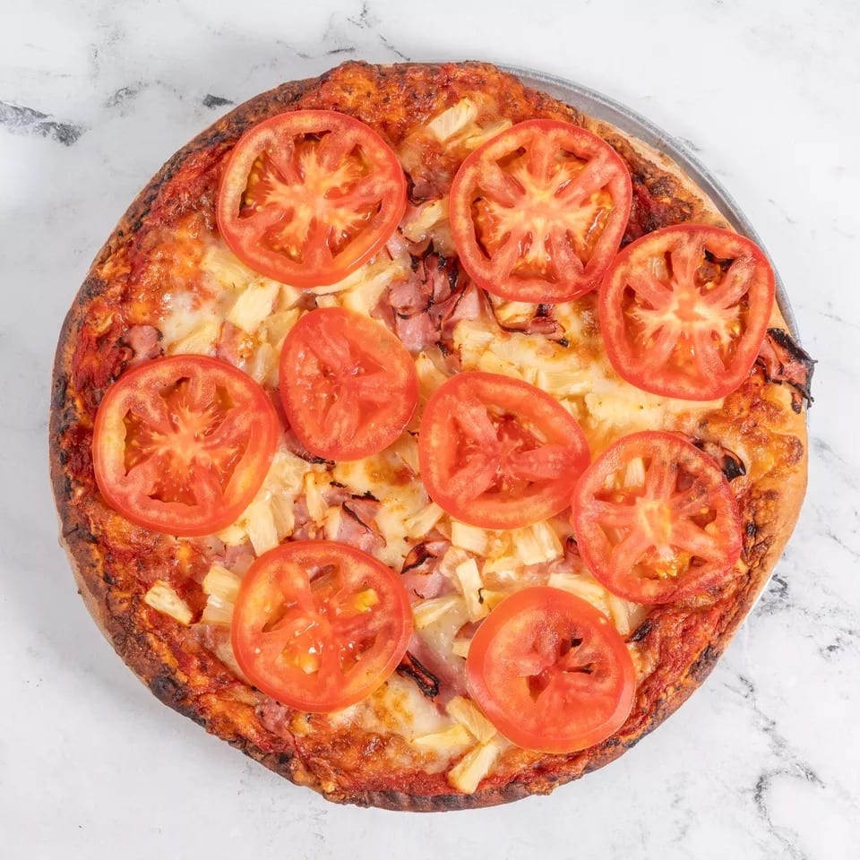 A whole pizza topped with ham, pineapple, and sliced tomatoes on a white marble surface.