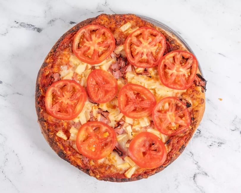 A whole pizza topped with ham, pineapple, and sliced tomatoes on a white marble surface.